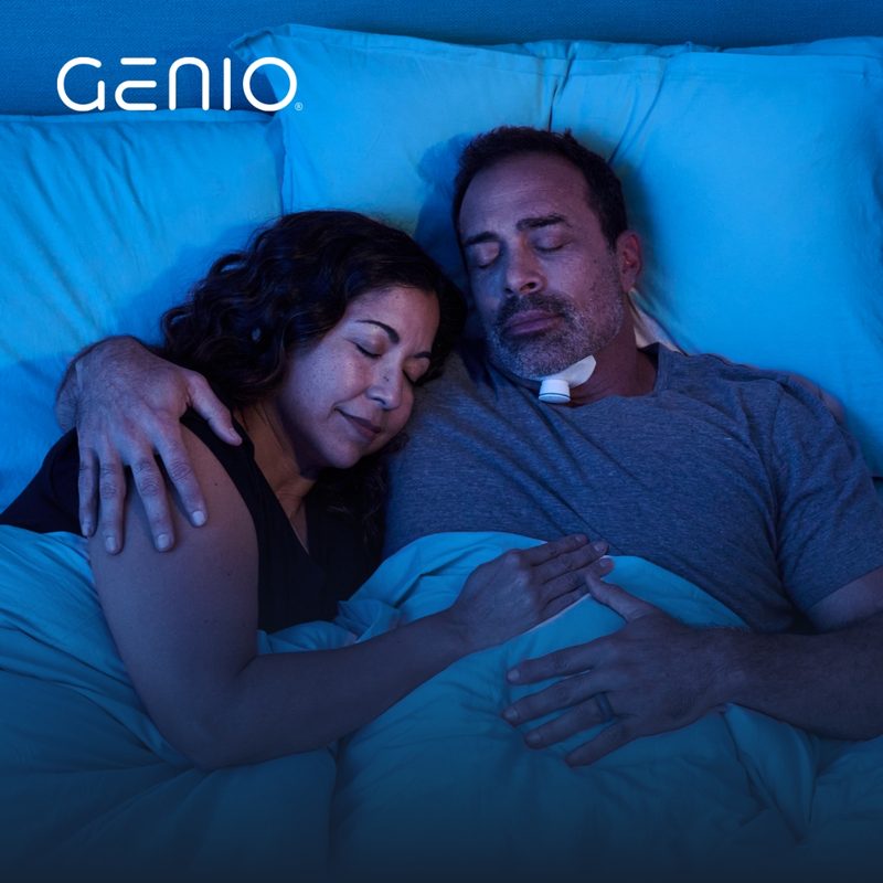 A man asleep in bed wearing the Genio activation chip on an adhesive patch under his chin, with his partner asleep beside him.